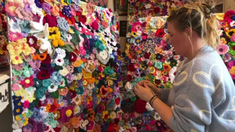 BBC Thousands of brightly coloured crochet flowers being pinned to a large pin boards. A woman is in the forefront working on attaching the flowers to the board. 