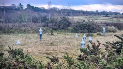 Sheryl Bishop A rural landscape with grassy fields and shrubs under a cloudy blue sky. Scattered across the field are several pale, human-shaped sculptures, some standing upright and others partially submerged in the ground.