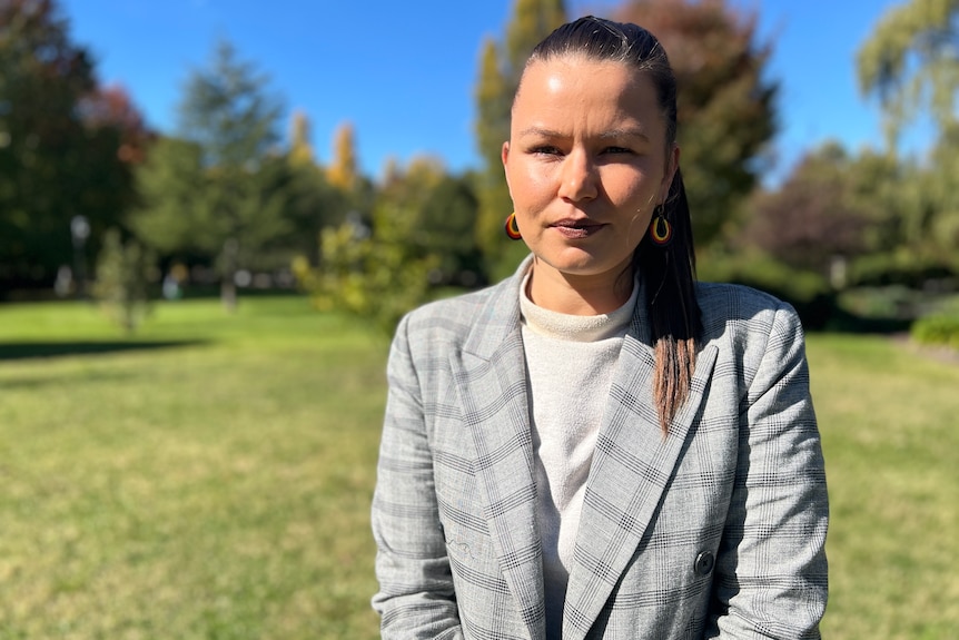 An Indigenous woman in a plaid suit jacket stands in a field looking serious.