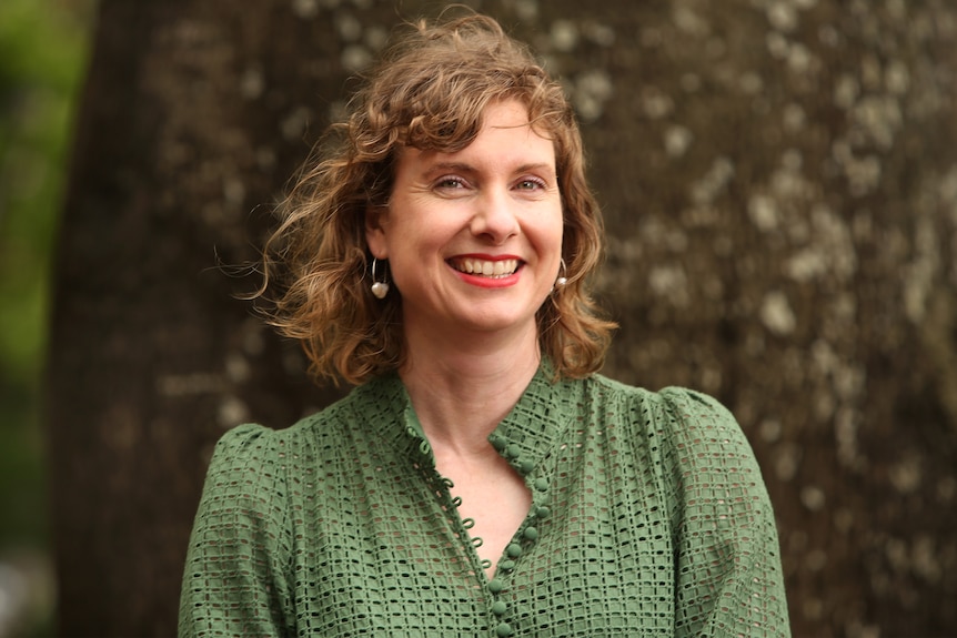 Writer Clare Fletcher smiles, she's standing in front of a tree, wearing a green blouse, with short bronde hair