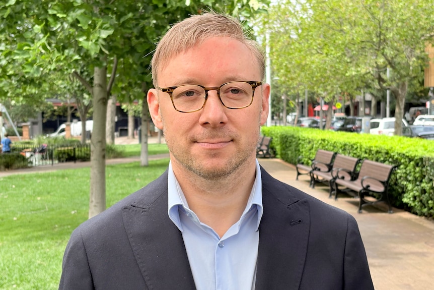 A person in a suit wearing glasses stands in a park smiling at the camera.