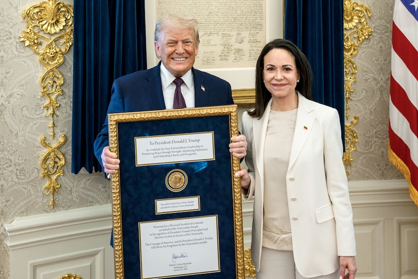 A man in a suit holds a frame with a medal inside, smiling, as a woman stands beside him