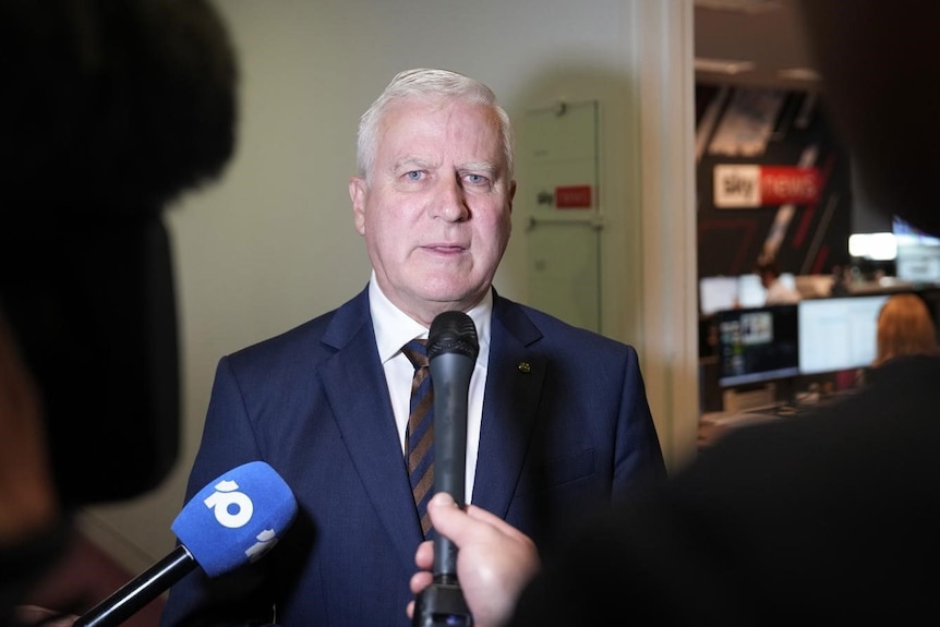 Michael McCormack in a suit in a hallway speaking into microphones