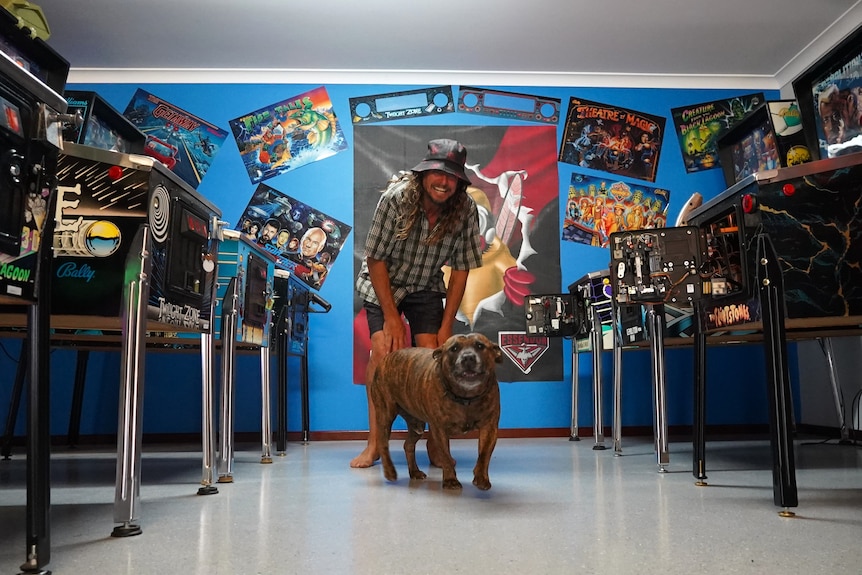 Azza Williamson smiles near a dog in a room full of pinball machines. 