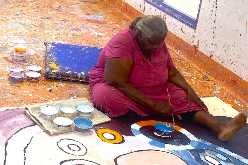 A woman in a pink dress sits on the ground, one leg out and paints a canvas on the floor using a stick. 
