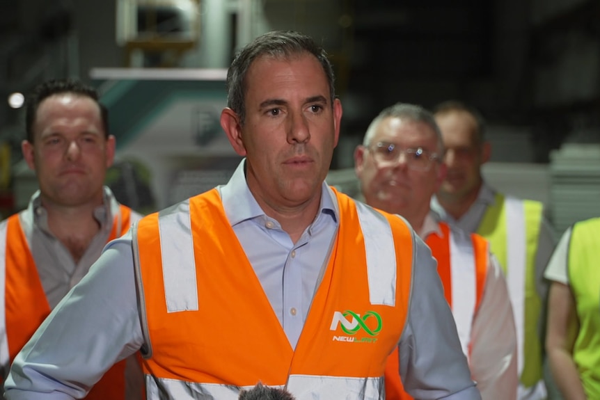 Chalmers, wearing an orange high-vis vest, speaks at a factory. Others, including Watt, stand behind also in high vis.