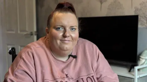 BBC Emily Salter, who is wearing a pink jumper, is in her wheelchair in her living room. There is a television on a cabinet and patterned wallpaper in the background. She is smiling at the camera. 