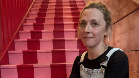 Pennie McKenzie sitting on red and pink painted stairs. Pennie has blonde hair which is in a bun and is wearing a black top with blue dungarees. She has big hoop earrings on and is looking at the camera. 
