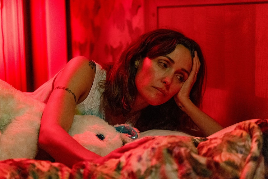 Rose Bryne lays on a bed with a stuffed toy in a red washed room