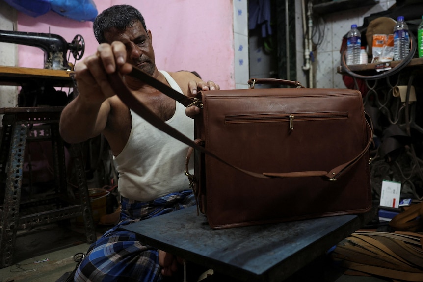 A man adjusts a brown leather strap on a bag that is sitting on a table in an inside workshop space