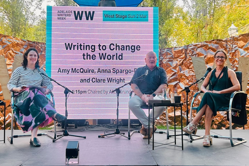 On an outdoor stage in front of a screen sits Alice Grundy, Jonathan Green and Clare Wright, behind microphones.