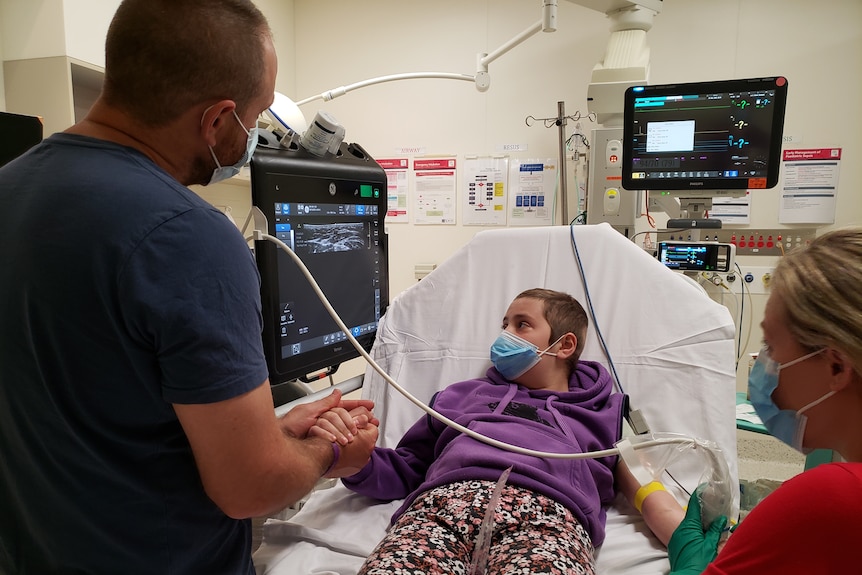 Abbey Barrett lies on a hospital bed looking at a monitor beside her while two adults hold her hands.