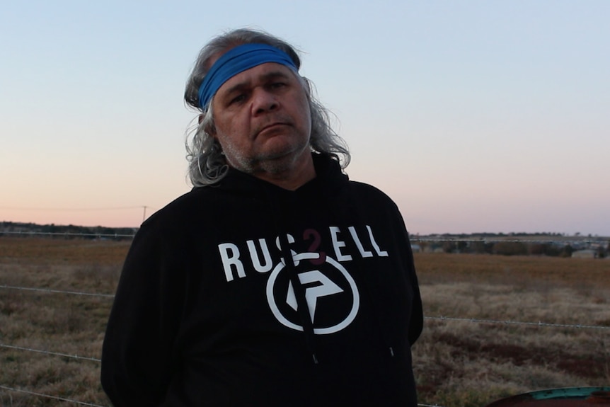A man wearing a headband stands in front of a field at dusk, looking serious