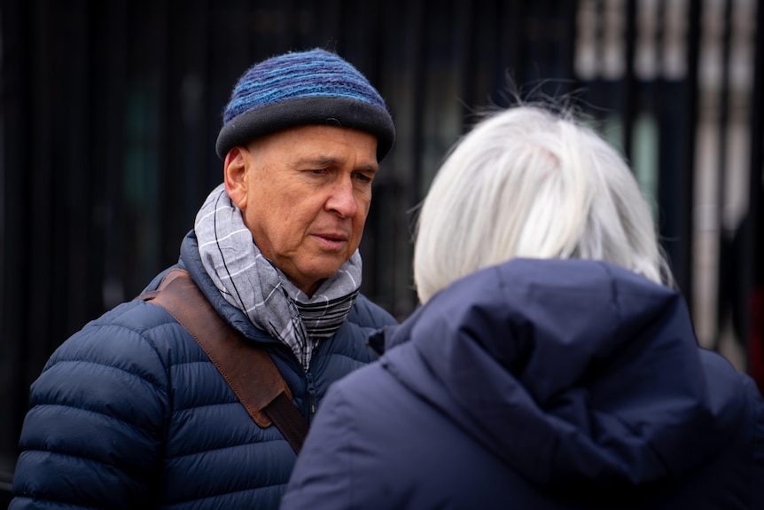 A man in a beanie and a puffer jacket speaks to a white-haired woman on a street.