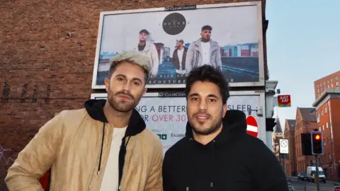 The Couture Club Ross Worswick and Scott Shashua are standing in front of a large billboard advertising The Couture Club. 