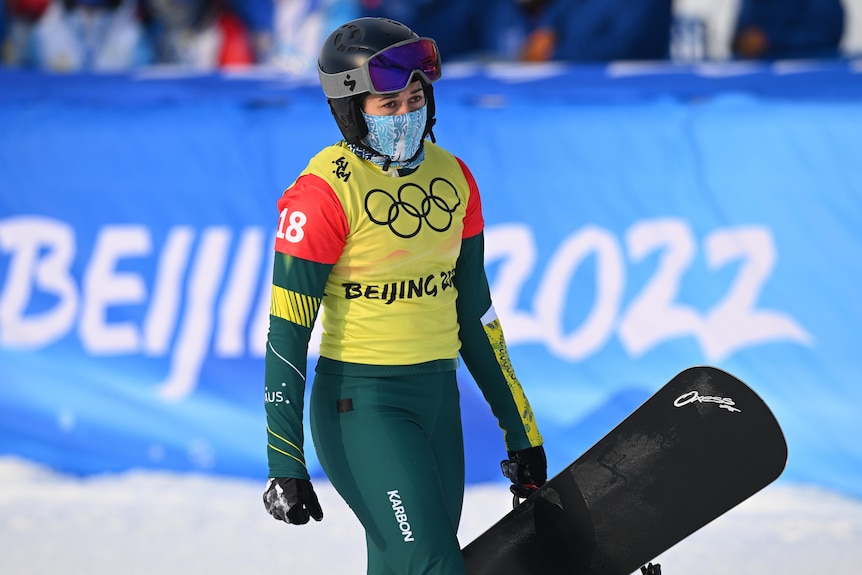 An Australian female snowboard cross competitor carries her snowboard at the 2022 Winter Olympics.