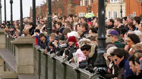 AFP via Getty Images Hundreds of people are packed behind the fence beside the Thames in west London. Many are holdings cameras