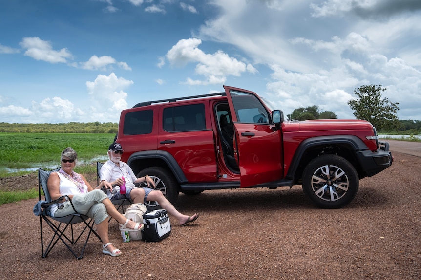 A couple sits beside a red four-wheel-drive on a dirt road