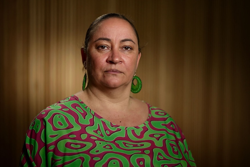 Image shows a woman with her hair pulled back. She is looking straight ahead and not smiling. She is wearing green earrings.