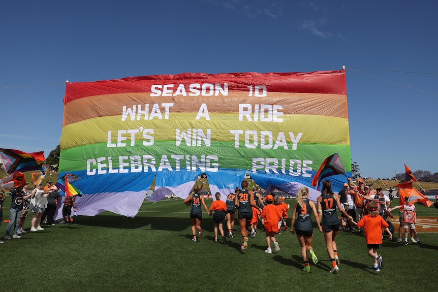 A rainbow banner reads 'season 10 what a ride let's win today celebrating pride' as AFLW players run through.