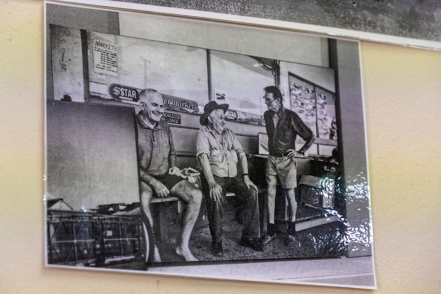 A black and white image of three men on a bench outside a supermarkey.
