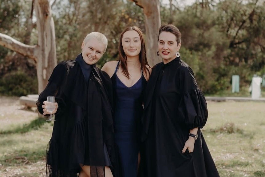 Three women stand outside with their arms around each other. One has a drink in her hand.