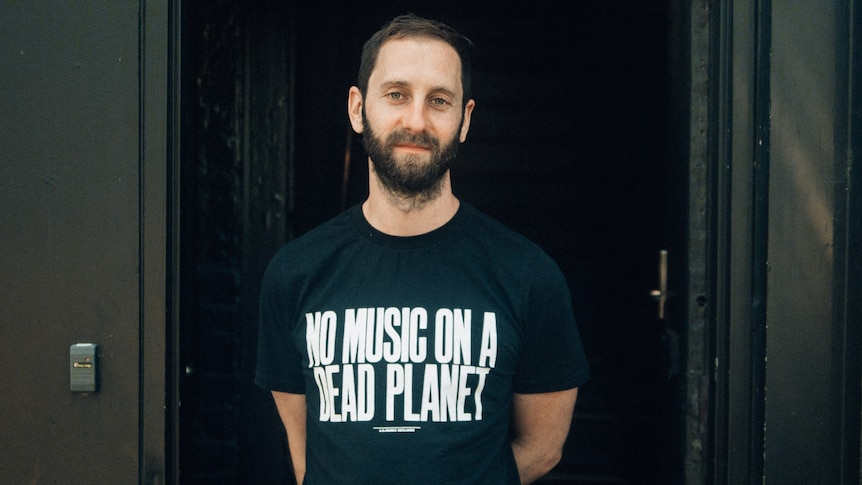 a white man with facial hair, wearing a black t-shirt that says 'no music on a dead planet'