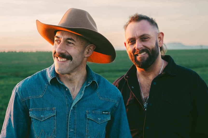 two men smiling in a field, one wears a cowboy hat and the other has a short beard.