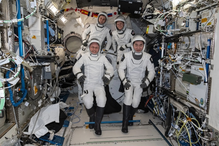 Four astronauts in spacesuits float inside the International Space Station, with electronics and cords in the background.