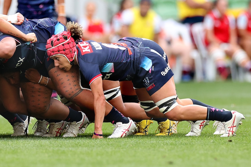 A woman in navy shorts and shirt and red headgear leans into a scrum.