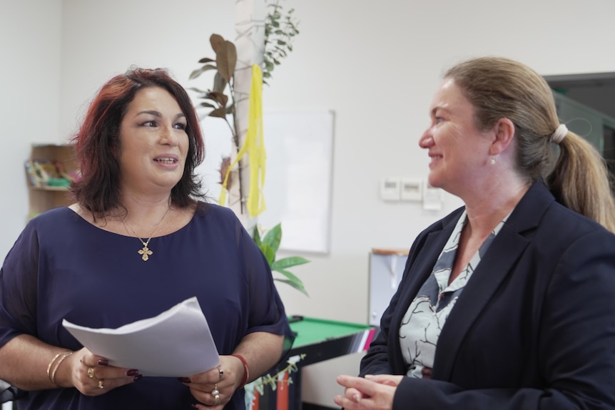 Two women talking, one with papers in hand, in education setting.