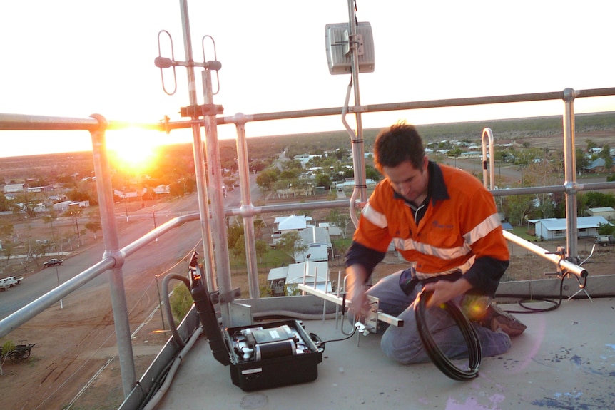 A man in high-vis handles an antenna while working on a roof above a country town.