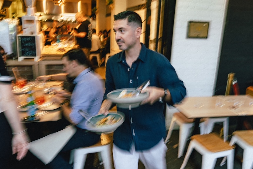 A man smiling but looking busy walks out of his restaurant to serve Italian dishes to patrons at outside tables.
