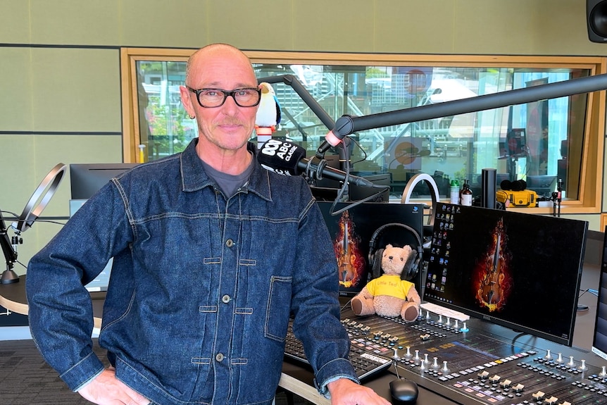Ed stands leaning one arm on the ABC Classic studio desk, he smiles to camera. 