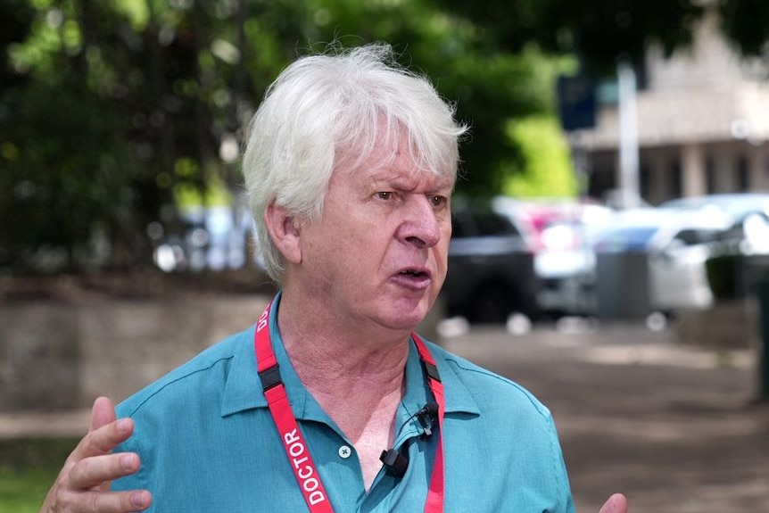 A doctor with grey hair and wearing a health lanyard, speaks outside a hospital.