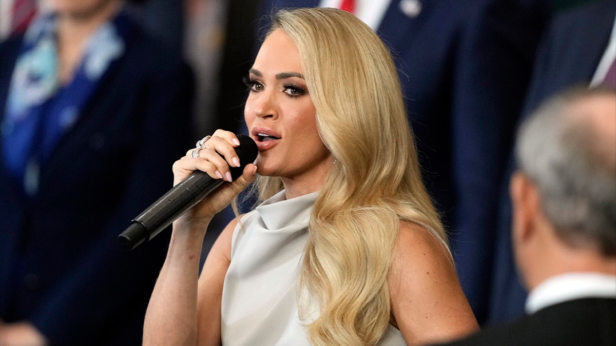 Carrie Underwood in a grey dress holds the microphone close to her mouth as she performs at the inauguration