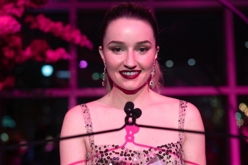 Kaitlyn Dever as Belle Gibson in Apple Cider Vinegar, smiling on a stage, about to give a speech.