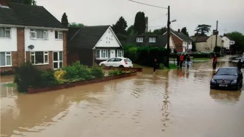 Gary Manning A residential street is heavily flooded, with muddy water covering the road and reaching up to the front gardens of houses. Several people stand on the higher, drier parts of the street observing the scene, while cars are partially submerged in the floodwater.