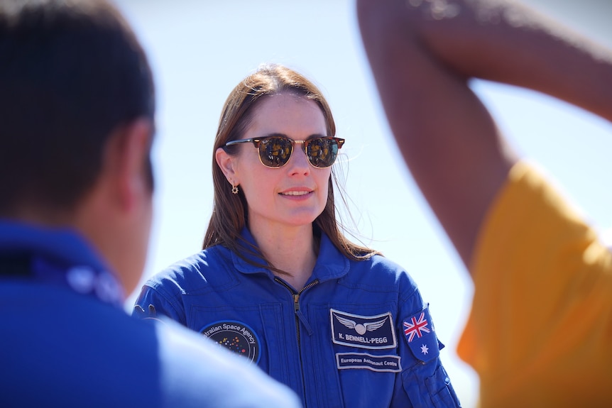 A brunette woman wears sunglasses and stands in front of two school kids with their backs to the camera.
