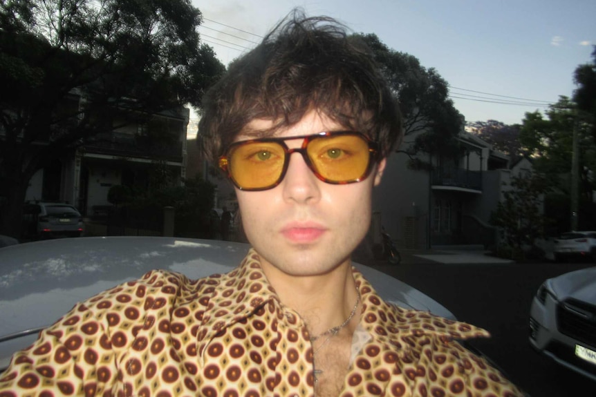 A man in a leopard print collared shirt and matching sunglasses casually takes a selfie with cars and houses behind him.