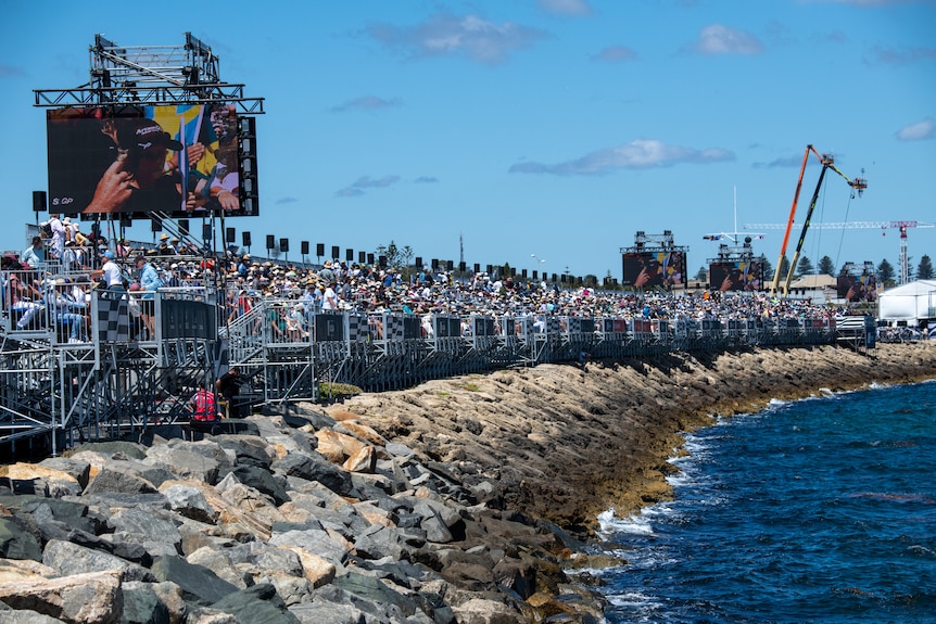 A large crowd gathered in a grandstand near a harbour. 
