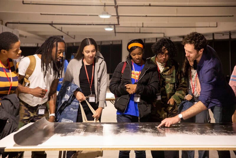 A group of six people, some wearing name badges, stand around a table examining a large black-and-white print while one man points and explains something to the others.