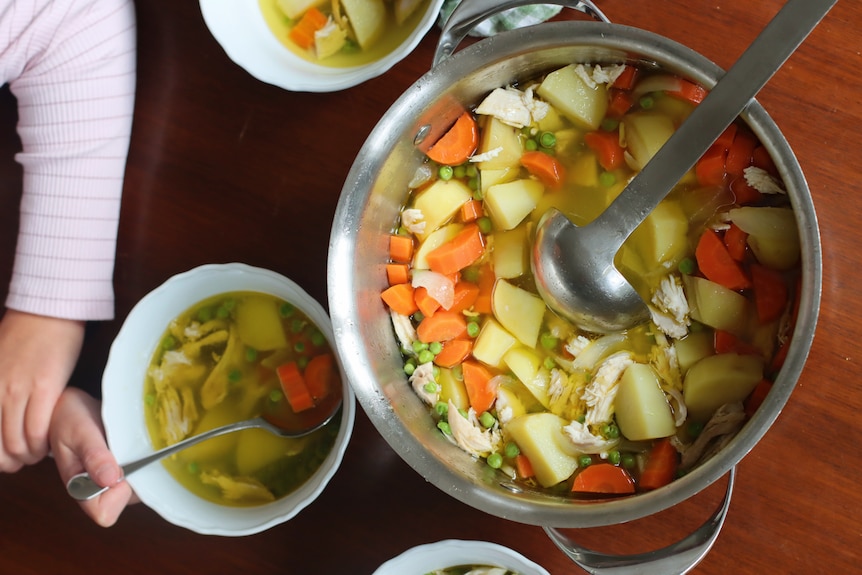 A stock pot of homemade chicken and vegetable soup with a ladle. Three bowls of soup sit alongside with a girl eating one