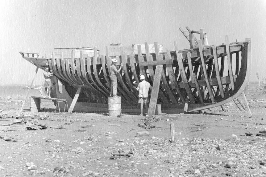 Archival image, black and white of a wooden boat being built, only the scaffolding so far, with two men working on it.