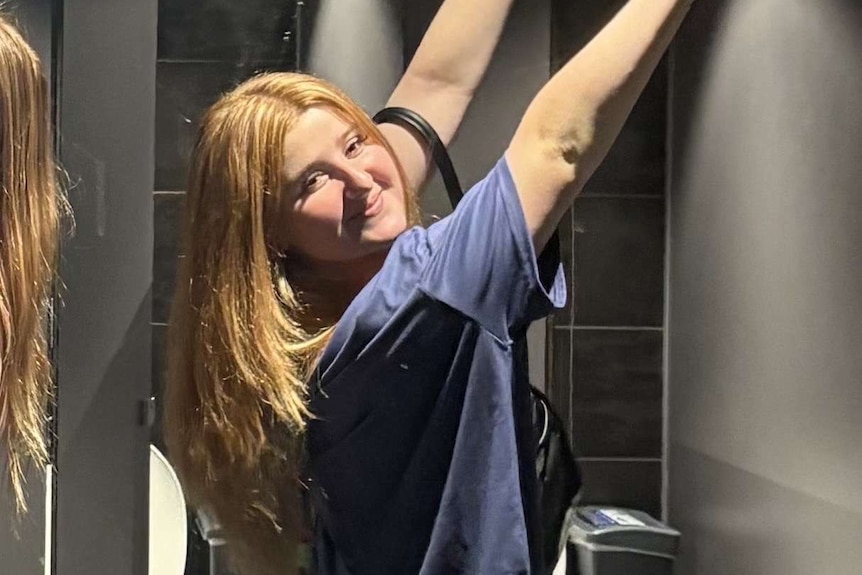 A mid-shot of a young woman with red hair smiling with her arms held straight up in the air in a public bathroom.