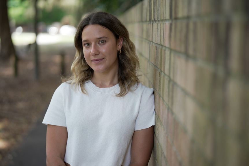 A young woman with shoulder length blond-brown hair and a white tshirt leans on a brick wall in a park.