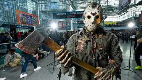 Getty Images A person dressed as Jason Voorheen - a character from the Friday the 13th movie series - is holding an axe and is wearing a hockey mask with a ripped top. Behind them, there is a crowd of people walking through a building.