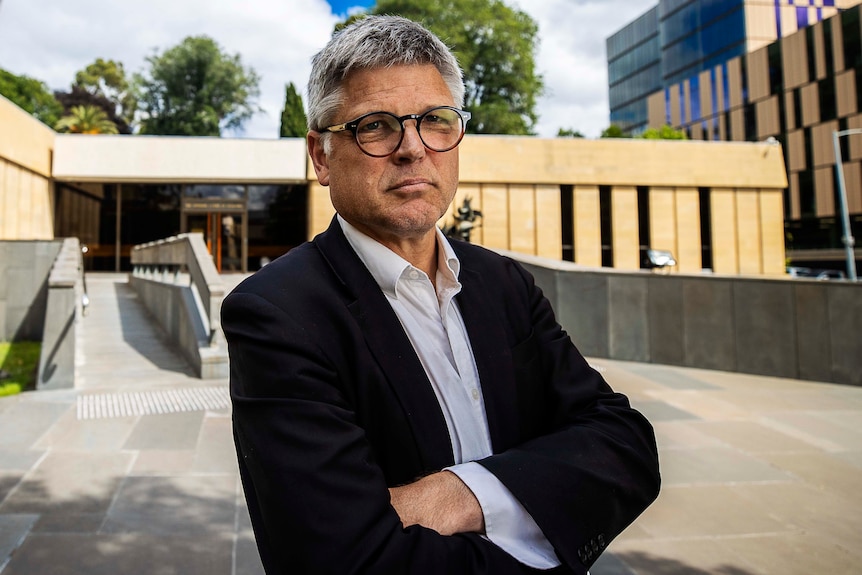 A man with silver hair and black rimmed glasses stands in front of a sandstone building