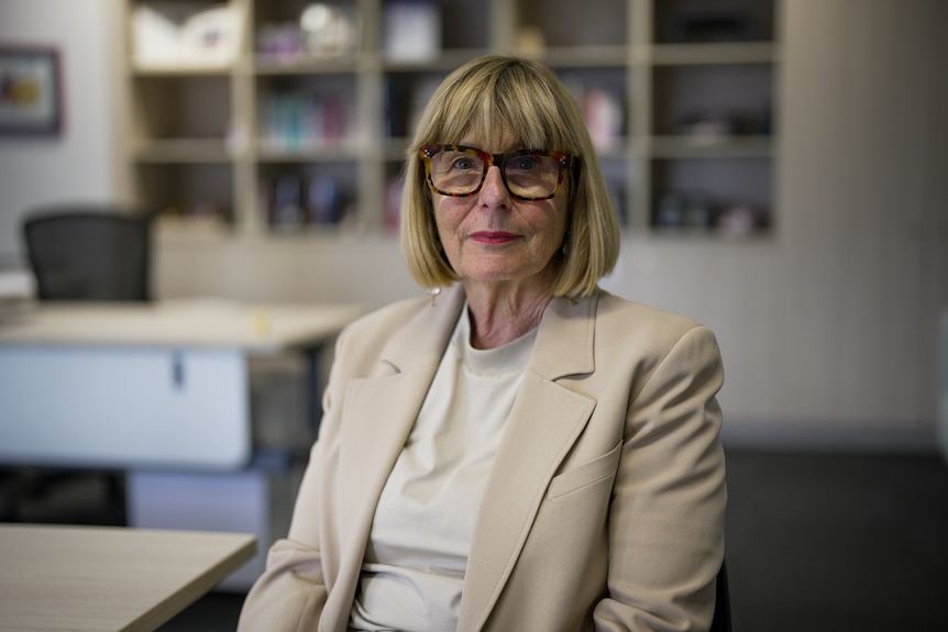 A woman with short blonde hair wearing a beige top and blazer sitting in an office.