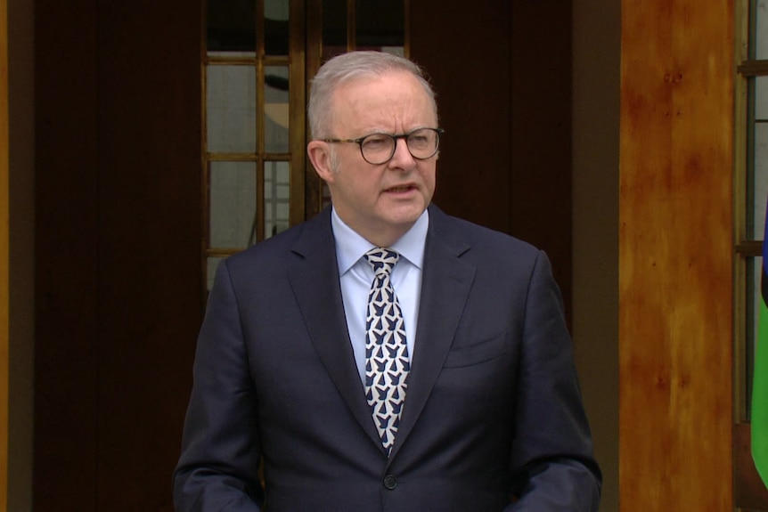 Albanese, wearing a suit, speaks from the doorway of Parliament House. A Torres Strait Islander flag can be seen to the right